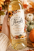 A hand holds a personalized white wine bottle with a gold foil cap and a fall-themed 'Give Thanks Happy Thanksgiving' label. The background is a cozy autumn setting with sunflowers, pumpkins, and warm bokeh lights, and a badge in the corner says 'PERFECT FOR HOSTESS GIFTS!'.