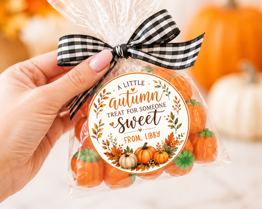 A hand holds a clear cellophane bag filled with small orange pumpkin-shaped treats, tied with a black-and-white gingham ribbon and a round autumn-themed label. The background is softly blurred with pumpkins and warm bokeh lights.