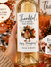 A hand holds a wine bottle decorated with a large, personalized Thanksgiving-themed label. The label features a cartoon turkey and the text 'Thankful for a family that drinks Happy Thanksgiving' with the names 'Reese & Mandy'.
