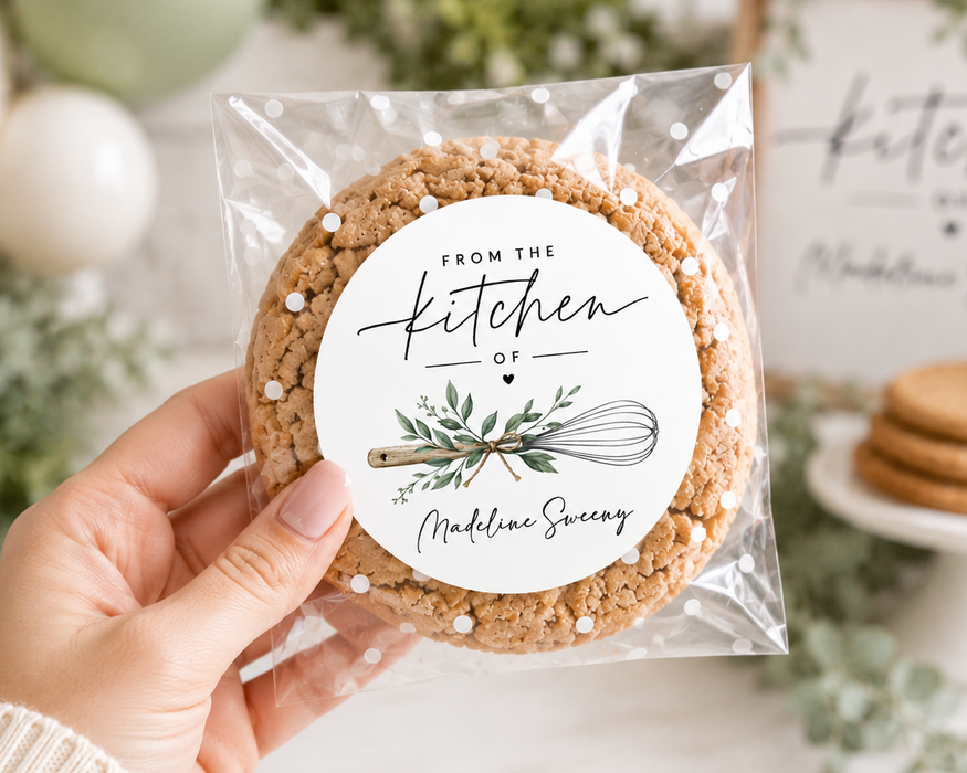 A hand holds a clear plastic bag containing a round oatmeal cookie with a white circular label that reads 'FROM THE kitchen OF Madison Sweetery.' The background is softly blurred with greenery, white balloons, and more cookies on a white stand.