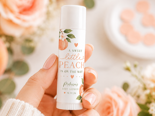 A hand holds a white lip balm tube labeled 'A Sweet Little Peach Is On The Way' with peach and green floral illustrations. The background is a soft, warm, blurred floral setting with peach roses and a white plate of peach candies.