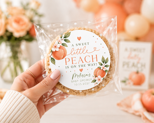 A hand holds a packaged round cookie with a peach-themed printed label that reads, 'A Sweet little PEACH is on the way! Melissa's Baby Shower 10.13.