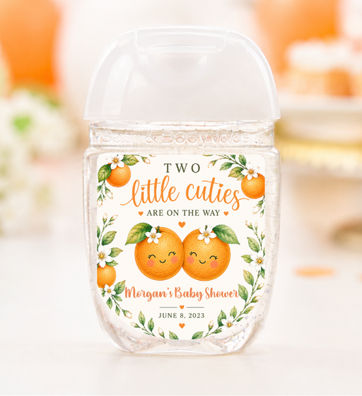 A clear plastic hand sanitizer bottle with a clear cap, decorated with an orange and green illustration of two smiling oranges and the text 'TWO little cuties ARE ON THE WAY Morgan's Baby Shower JUNE 8, 2023'. The background is a soft-focus white and orange lifestyle setting.