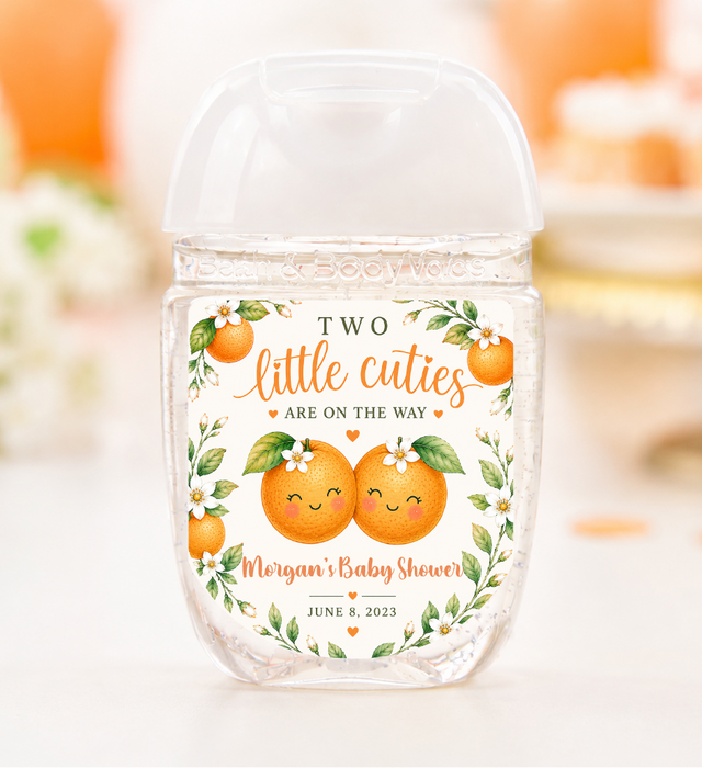 A clear plastic hand sanitizer bottle with a clear cap, decorated with an orange and green illustration of two smiling oranges and the text 'TWO little cuties ARE ON THE WAY Morgan's Baby Shower JUNE 8, 2023'. The background is a soft-focus white and orange lifestyle setting.
