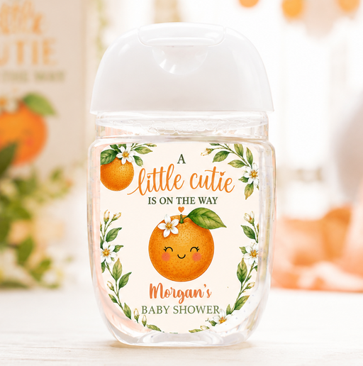 A clear plastic hand sanitizer bottle with a white cap, decorated with a custom orange-themed label that reads 'A little cutie is on the way' and 'Morgan's Baby Shower'. The background is a soft-focus, warm-toned autumn setting with pumpkins and a matching blurred sign.