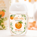 A clear plastic hand sanitizer bottle with a white cap, decorated with a custom orange-themed label that reads 'A little cutie is on the way' and 'Morgan's Baby Shower'. The background is a soft-focus, warm-toned autumn setting with pumpkins and a matching blurred sign.