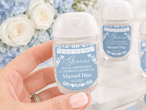A hand holds a small clear hand sanitizer bottle with a white cap and a blue personalized label. Two more matching bottles sit on a white marble surface, along with decorative blue lace, butterflies, flowers, and a small house figurine.