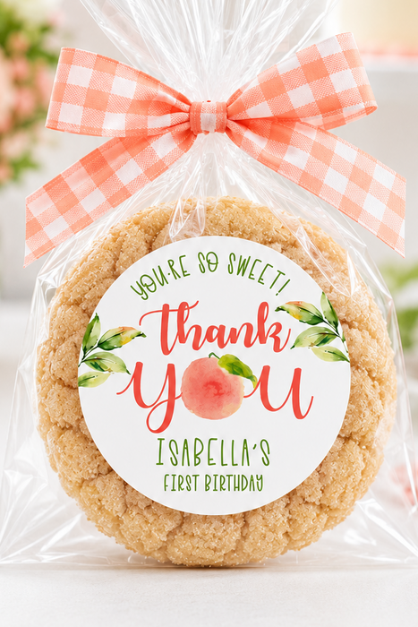 A round peach-themed cookie in a clear cellophane bag tied with a peach-and-white gingham ribbon, featuring a round 'thank you' label that reads 'Isabella's First Birthday' with a peach illustration. The background is a softly blurred party setting with pink balloons, flowers, and a cake.