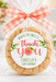 A round peach-themed cookie in a clear cellophane bag tied with a peach-and-white gingham ribbon, featuring a round 'thank you' label that reads 'Isabella's First Birthday' with a peach illustration. The background is a softly blurred party setting with pink balloons, flowers, and a cake.