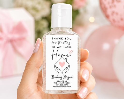 A hand with pink manicured nails holds a clear plastic hand sanitizer bottle with a white cap and a custom white label. The background is a soft-focus pink-themed setting with balloons and a gift box.