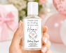 A hand with pink manicured nails holds a clear plastic hand sanitizer bottle with a white cap and a custom white label. The background is a soft-focus pink-themed setting with balloons and a gift box.