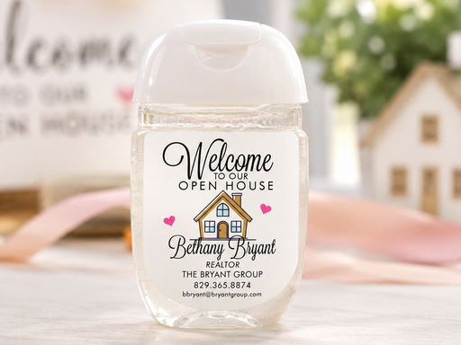 A personalized hand sanitizer bottle with a clear cap and a custom printed label reading 'Welcome to our OPEN HOUSE Bethany Bryant' on a light marble surface.