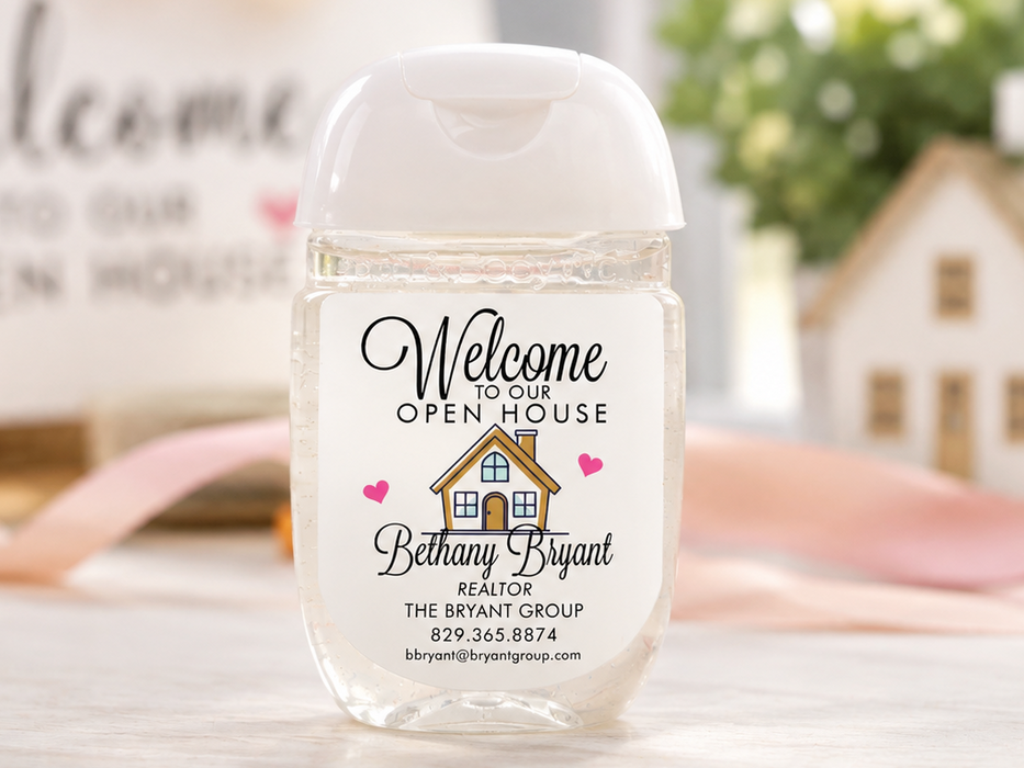 A personalized hand sanitizer bottle with a clear cap and a custom printed label reading 'Welcome to our OPEN HOUSE Bethany Bryant' on a light marble surface.