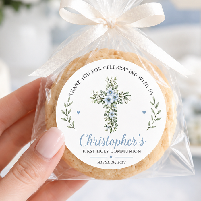 A hand holds a round decorated cookie sealed in a clear cellophane bag tied with a cream ribbon. The front label reads 'Thank you for celebrating with us Christopher's First Holy Communion April 10, 2024' with a floral cross illustration.