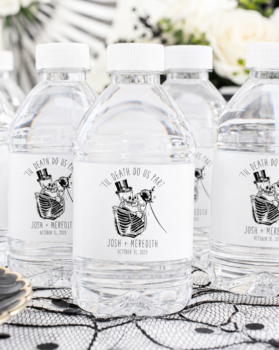 Five clear plastic water bottles with white caps arranged on a black-and-white patterned tablecloth. Each bottle has a white label featuring a black skeleton illustration wearing a top hat and bow tie, with the text 'THE DEAD DO NO HARM' and personalized names and date.
