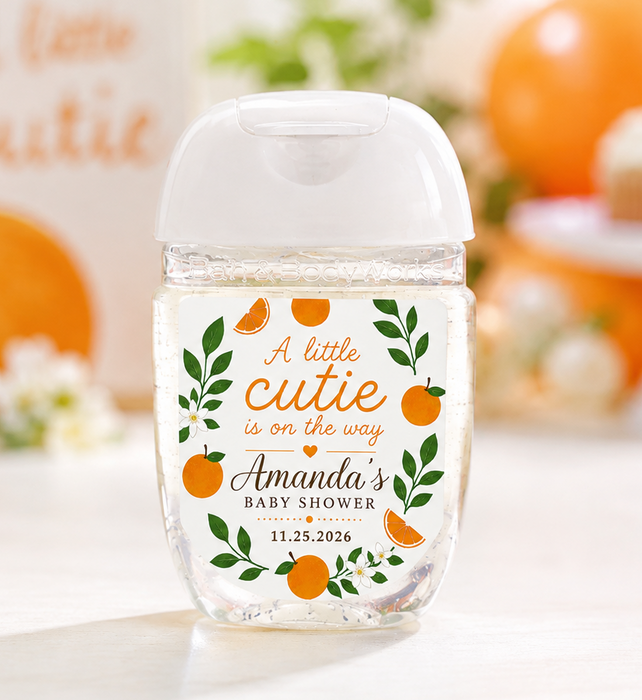 A clear plastic hand sanitizer bottle with a white flip-top cap, decorated with an orange and green 'A little cutie is on the way Amanda's baby shower' label. The bottle is set on a white surface with a warm, blurred background of pumpkins, cupcakes, and a sign reading 'A little Cutie'.