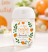 A clear plastic hand sanitizer bottle with a white flip-top cap, decorated with an orange and green 'A little cutie is on the way Amanda's baby shower' label. The bottle is set on a white surface with a warm, blurred background of pumpkins, cupcakes, and a sign reading 'A little Cutie'.