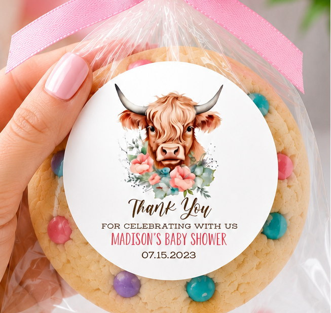 A hand holds a round decorated cookie wrapped in clear cellophane and tied with a pink ribbon. The cookie has a colorful cow illustration and personalized text on the label.