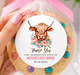 A hand holds a round decorated cookie wrapped in clear cellophane and tied with a pink ribbon. The cookie has a colorful cow illustration and personalized text on the label.