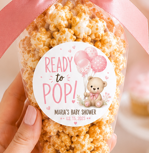 A hand holds a clear plastic cone filled with caramel popcorn and topped with a pink satin bow and a round 'Ready to Pop! Wrigley's Baby Shower' label. The background is a soft pink baby shower theme with balloons, cupcakes, and a teddy bear.