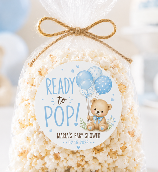 A clear cellophane bag filled with popcorn, tied with a twine bow, and featuring a round custom label that reads 'READY to POP! MARIA'S BABY SHOWER' with a teddy bear and balloons, set against a softly blurred blue and white party background.