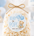 A clear cellophane bag filled with popcorn, tied with a twine bow, and featuring a round custom label that reads 'READY to POP! MARIA'S BABY SHOWER' with a teddy bear and balloons, set against a softly blurred blue and white party background.