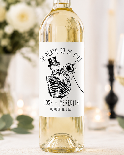A bottle of white wine with a custom skeleton wedding label reading 'TIL DEATH DO US PART' and 'JOSH + MEREDITH OCTOBER 31, 2023', set on a table with a blurred wine glass, flowers, and candles in the background.