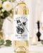 A bottle of white wine with a custom skeleton wedding label reading 'TIL DEATH DO US PART' and 'JOSH + MEREDITH OCTOBER 31, 2023', set on a table with a blurred wine glass, flowers, and candles in the background.