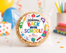 A round sugar cookie sealed in a clear plastic bag with a colorful circular 'Welcome Back to School!' label. The label includes school-themed illustrations and the text 'FROM: MRS.