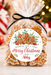 A clear cellophane bag filled with gingerbread cookies, tied with a black ribbon, and featuring a round holiday label that reads 'Wishing You a Very Merry Christmas From Abby.' The background shows a softly blurred Christmas scene with warm lights and ornaments.