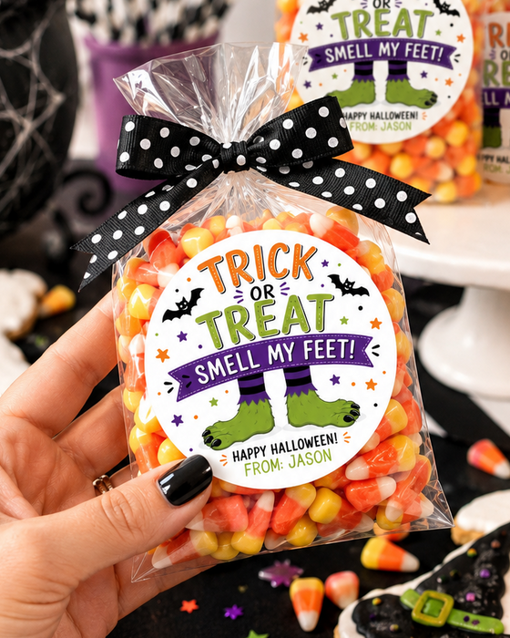 A hand holds a clear bag of Halloween candy with a black polka-dot bow and a round label reading 'Trick or Treat Smell My Feet! Happy Halloween from Jess.' Other similar candy bags and Halloween decorations are visible on a white pedestal and around the table.