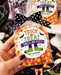 A hand holds a clear bag of Halloween candy with a black polka-dot bow and a round label reading 'Trick or Treat Smell My Feet! Happy Halloween from Jess.' Other similar candy bags and Halloween decorations are visible on a white pedestal and around the table.