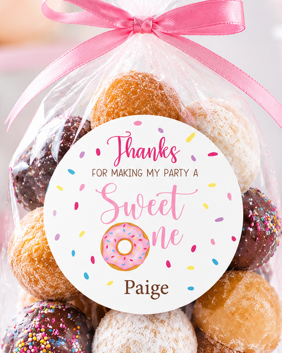 A clear cellophane gift bag filled with assorted donut-shaped treats tied with a pink ribbon and topped with a round printed tag that reads 'Thanks for making my party a Sweet one Paige.' The bag is photographed on a light pink party-themed surface with blurred pink flowers and confetti in the background.