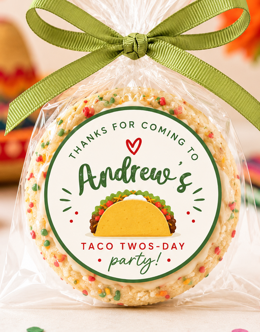A round decorated cookie or candy cookie wrapped in clear cellophane and tied with an olive green ribbon, featuring a custom printed round label that reads 'Thanks for coming to Andrew's Taco Twos-Day party!' with a taco illustration. The photo is set in a festive, blurred Mexican-themed party environment with papel picado, a sombrero, and flowers.