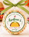 A round decorated cookie or candy cookie wrapped in clear cellophane and tied with an olive green ribbon, featuring a custom printed round label that reads 'Thanks for coming to Andrew's Taco Twos-Day party!' with a taco illustration. The photo is set in a festive, blurred Mexican-themed party environment with papel picado, a sombrero, and flowers.