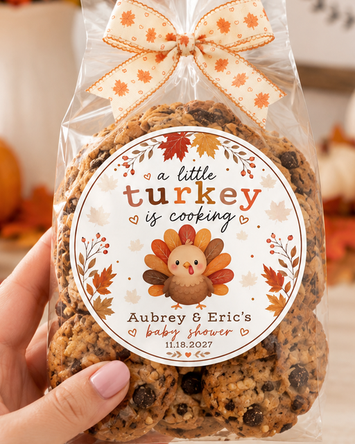 A hand holds a clear bag of personalized turkey-shaped cookies decorated with a fall-themed label and an orange patterned ribbon bow. The background is a warm, autumnal setting with pumpkins, fall leaves, and a 'gather' sign.