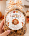 A hand holds a clear bag of personalized turkey-shaped cookies decorated with a fall-themed label and an orange patterned ribbon bow. The background is a warm, autumnal setting with pumpkins, fall leaves, and a 'gather' sign.