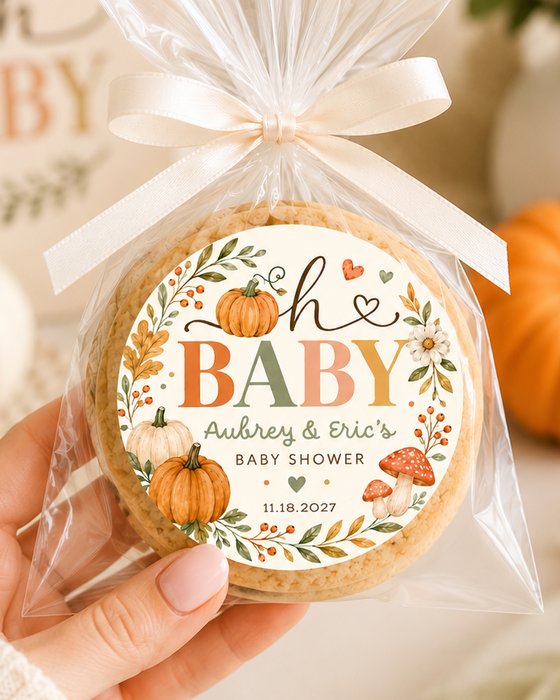 A hand holds a round baby shower favor cookie wrapped in clear cellophane and tied with a cream ribbon. The front has a decorative printed label reading 'oh BABY Aubrey & Eric's BABY SHOWER' with fall-themed illustrations.