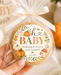 A hand holds a round baby shower favor cookie wrapped in clear cellophane and tied with a cream ribbon. The front has a decorative printed label reading 'oh BABY Aubrey & Eric's BABY SHOWER' with fall-themed illustrations.