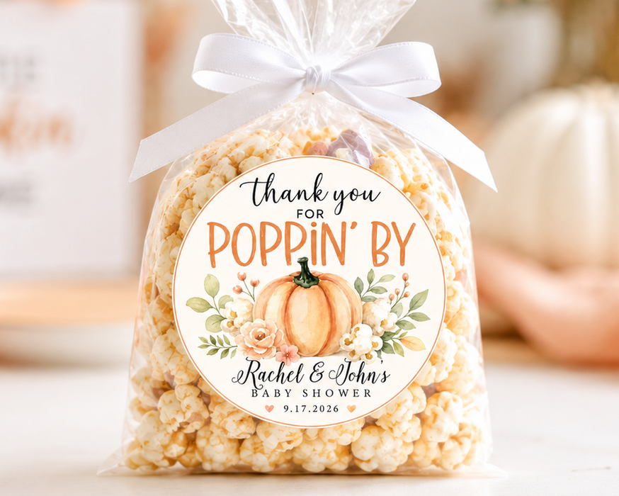 A clear cellophane bag filled with popcorn and tied with a white ribbon, featuring a round autumn-themed label that reads 'thank you for POPPIN' BY Rachel & John's Baby Shower 9.17.