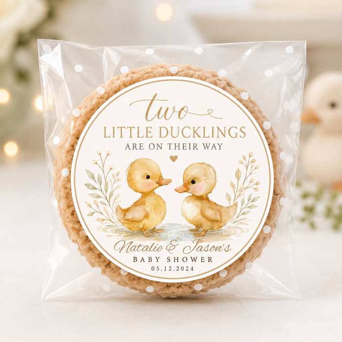 A round decorated cookie sealed in a clear plastic bag with a printed label that reads 'two LITTLE DUCKLINGS ARE ON THEIR WAY' and shows two yellow ducklings with the names 'Nicole & Jason's BABY SHOWER' and a date. The photo is taken on a light tabletop with soft blurred flowers and a duck toy in the background.