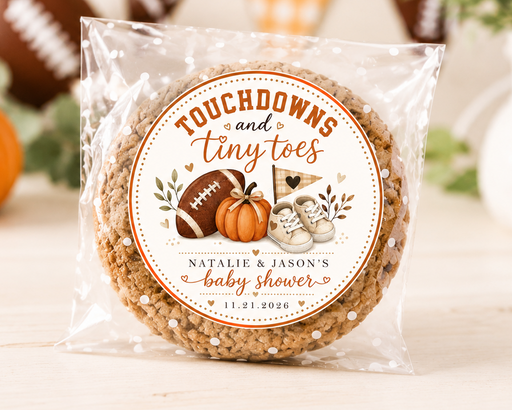 A packaged round cookie or candy with a custom football-themed label reading 'Touchdowns and Tiny toes' and 'Natalie & Jason's baby shower,' photographed on a light wood surface with pumpkins and blurred decor in the background.