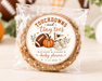 A packaged round cookie or candy with a custom football-themed label reading 'Touchdowns and Tiny toes' and 'Natalie & Jason's baby shower,' photographed on a light wood surface with pumpkins and blurred decor in the background.