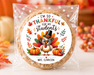 A round custom-printed cookie or biscuit sealed in a clear cellophane bag, placed on a white surface with autumn decorations in the background. The printed label features a turkey illustration and the text 'I'M SO THANKFUL FOR MY students' and 'MRS.