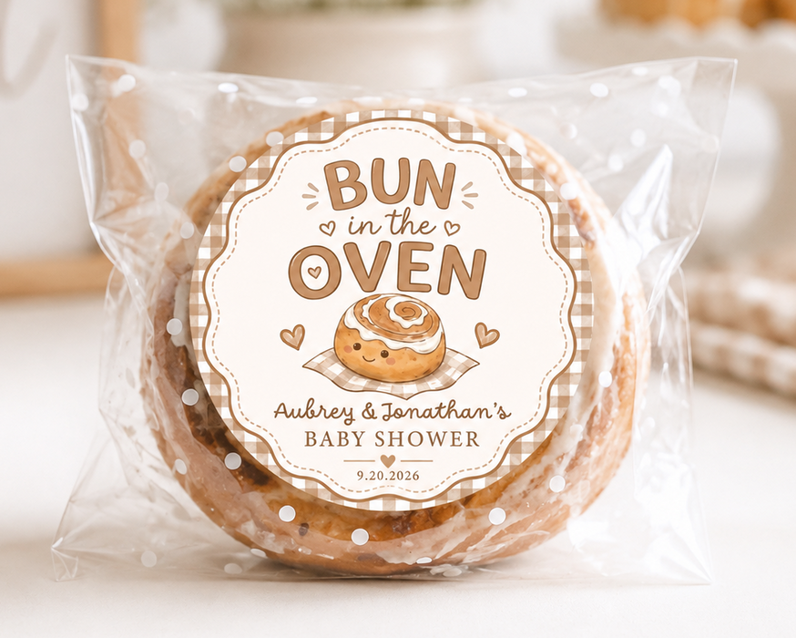 A round baked good wrapped in a clear plastic bag with a custom printed round label reading 'BUN in the OVEN' and 'Aubrey & Jonathan's Baby Shower,' shown on a light tabletop with a softly blurred background.