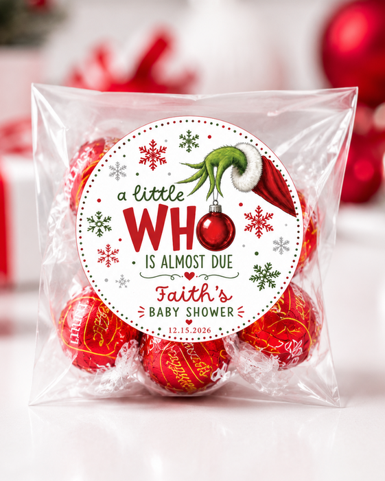 A clear plastic bag containing red wrapped baby shower favors with a round printed label that reads 'a little WHO IS ALMOST DUE Faith's BABY SHOWER' and features the Grinch graphic, photographed on a white surface with blurred red holiday ornaments in the background.