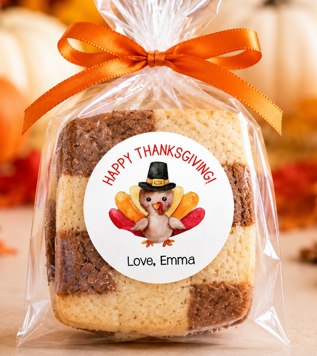 A square layered cookie or cake slice wrapped in a clear cellophane bag tied with an orange ribbon, featuring a round personalized Thanksgiving label with a turkey illustration and the text 'Happy Thanksgiving! Love, Emma.' The background shows autumn decor, including pumpkins, fall leaves, and warm blurred lights.