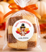 A square layered cookie or cake slice wrapped in a clear cellophane bag tied with an orange ribbon, featuring a round personalized Thanksgiving label with a turkey illustration and the text 'Happy Thanksgiving! Love, Emma.' The background shows autumn decor, including pumpkins, fall leaves, and warm blurred lights.