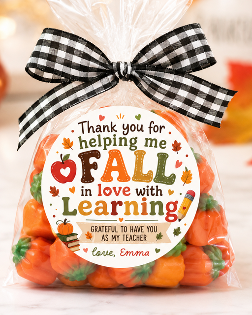 A clear cellophane gift bag filled with small orange pumpkin-shaped candies, tied with a black-and-white gingham bow. A round printed label on the front reads, 'Thank you for helping me FALL in love with Learning' with autumn-themed decorations.