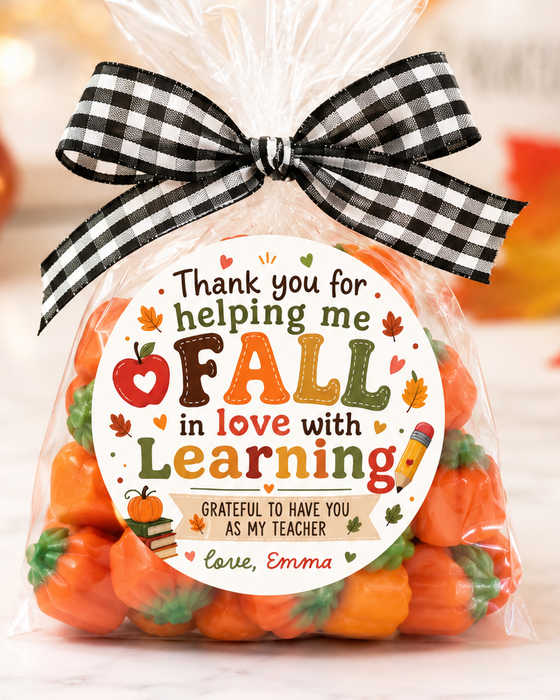 A clear cellophane gift bag filled with small orange pumpkin-shaped candies, tied with a black-and-white gingham bow. A round printed label on the front reads, 'Thank you for helping me FALL in love with Learning' with autumn-themed decorations.
